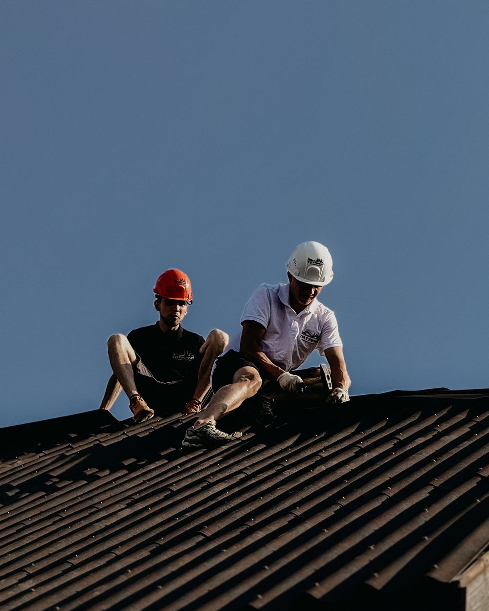 Two roofers in hard hats working on a metal roof under a clear sky